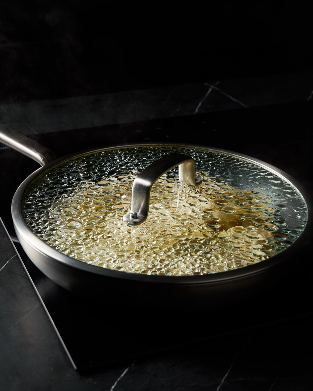 Non-stick frying pan with glass lid steaming on a black stovetop, kitchen cookware