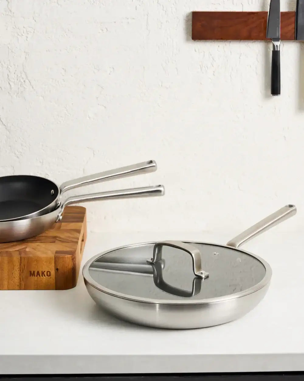 Mako stainless steel non-stick pans and glass lid on white countertop with wooden block and wall knives