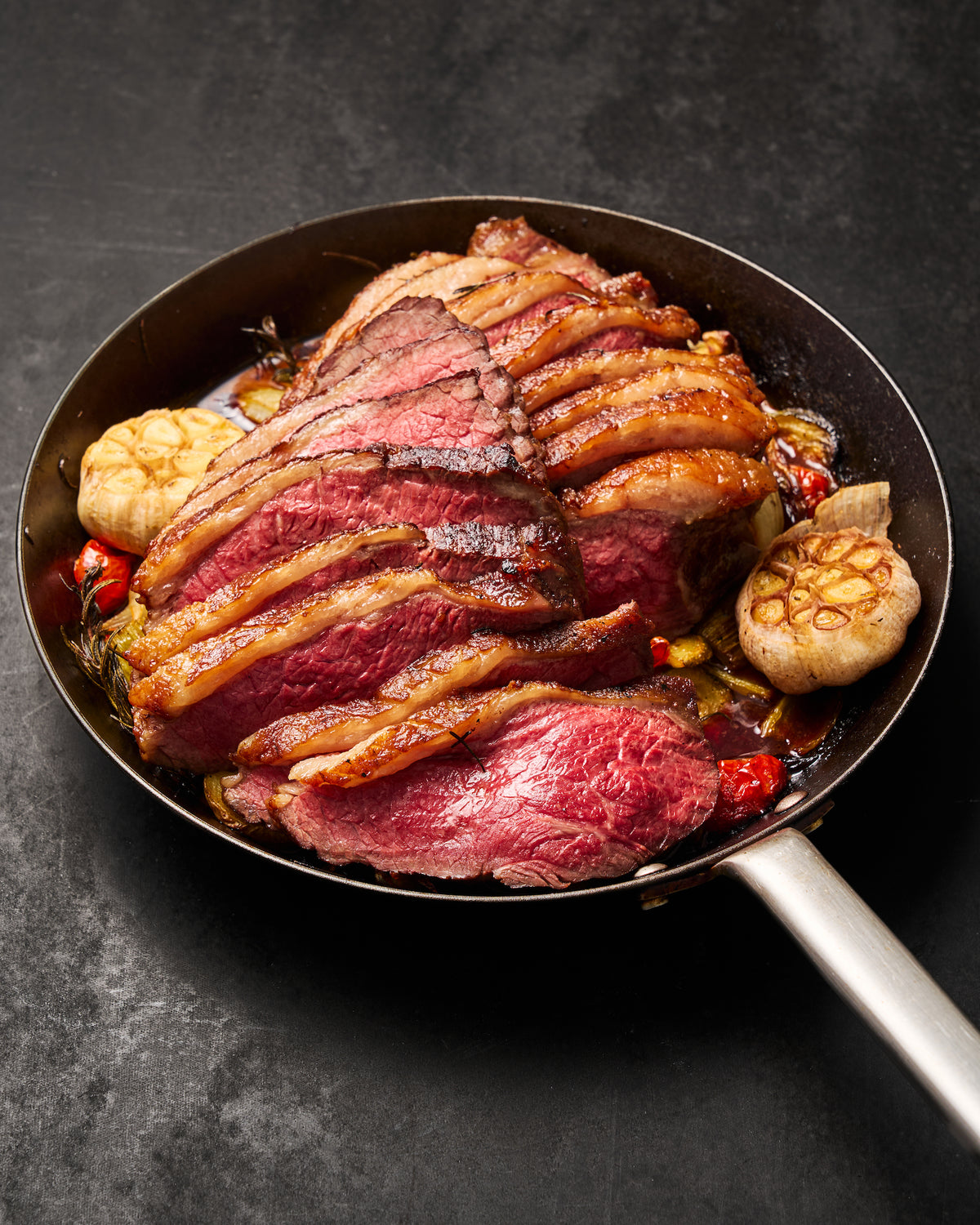 Sliced cooked beef steak with crispy browned strips, served in a dark frying pan with garlic, red peppers, and oil on a dark textured background.