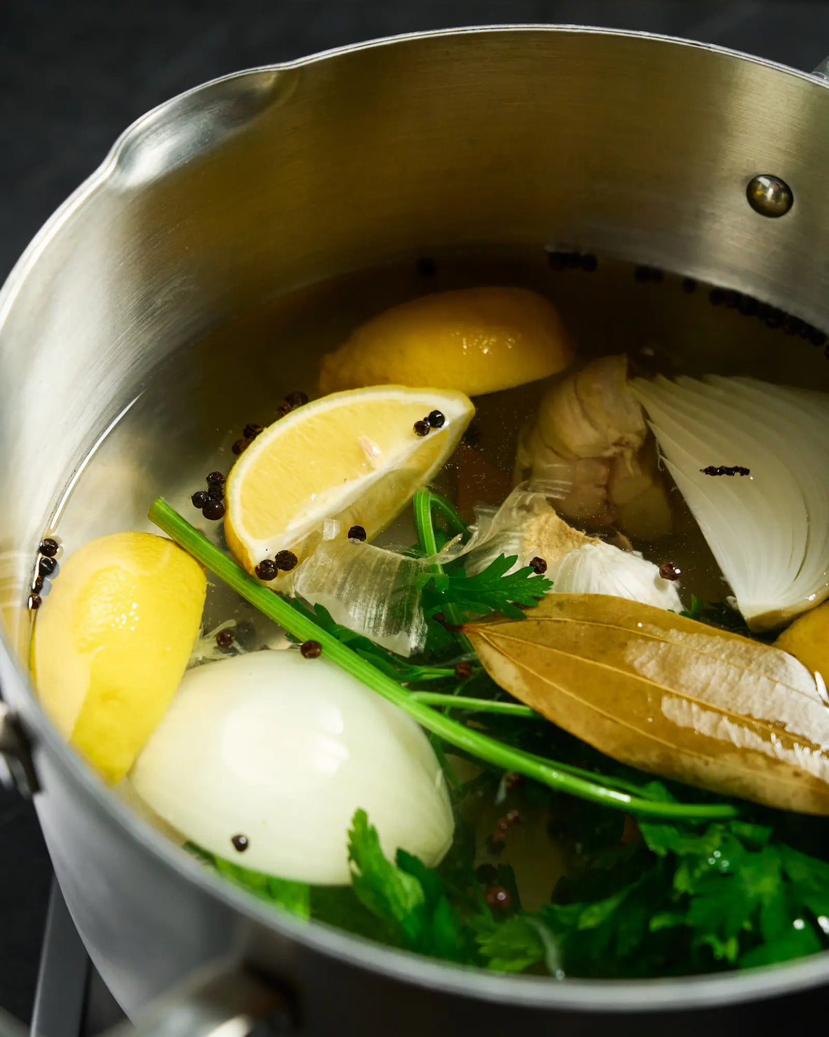 Lemon wedges, garlic, onion, bay leaves, peppercorns and fresh herbs soaking in chicken brine in a stainless steel saucepan.