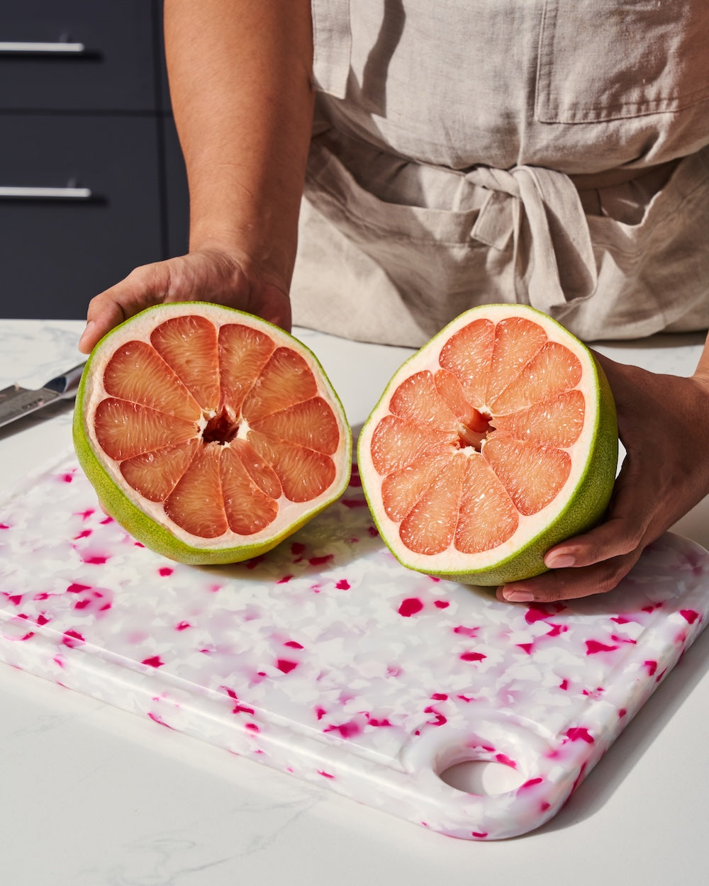Person holding halved pomelo on a white and pink cutting board in a kitchen setting