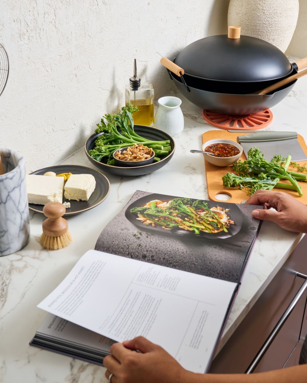 Modern kitchen scene with wok, cookbook, fresh broccolini, sauce, tofu, and walnuts on counter.