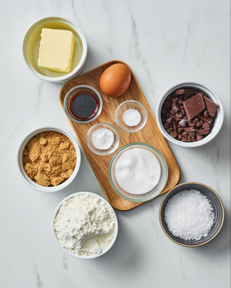 Overhead view of skillet cookie ingredients - butter, a brown egg, vanilla extract, baking soda, white sugar, brown sugar, flour, chocolate chunks and chips, and flaky sea salt.
