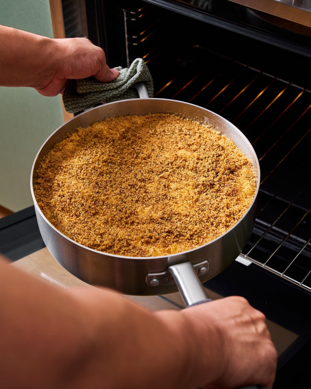 Person placing a stainless steel sauté pan with baked crumb topping into an oven