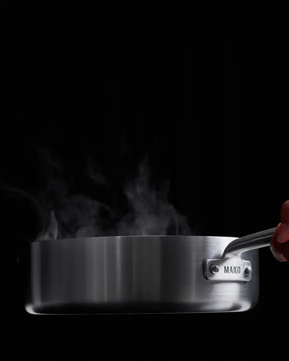 Steam rising from a stainless steel Mako sauté pan with a hand holding the handle, cooking action
