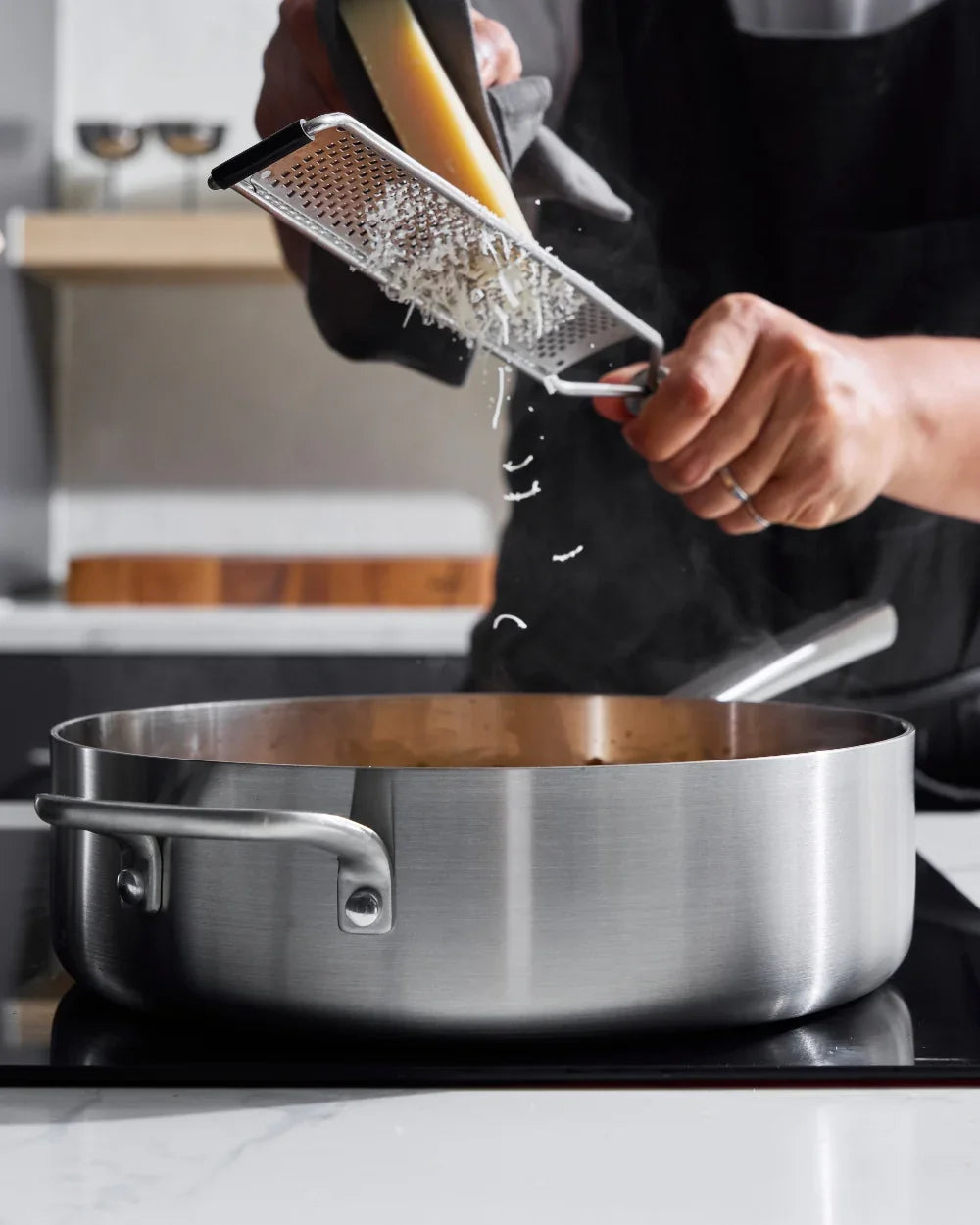 Hands grating cheese over a steaming stainless steel Mako saute pan in a modern kitchen
