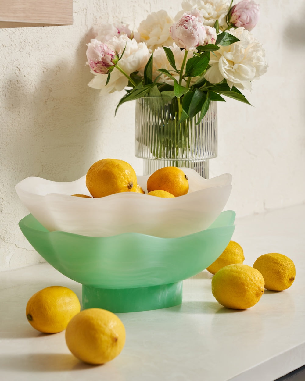 Modern glass fruit bowl with lemons beside vase of white and pink flowers on countertop