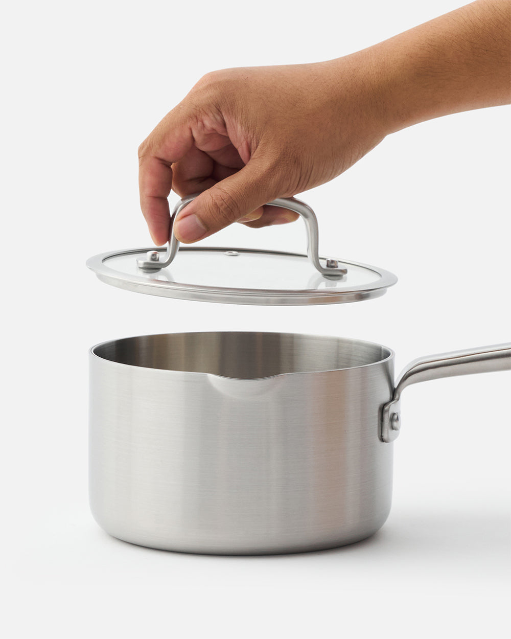 Hand lifting lid from stainless steel saucepan with handle on white background.