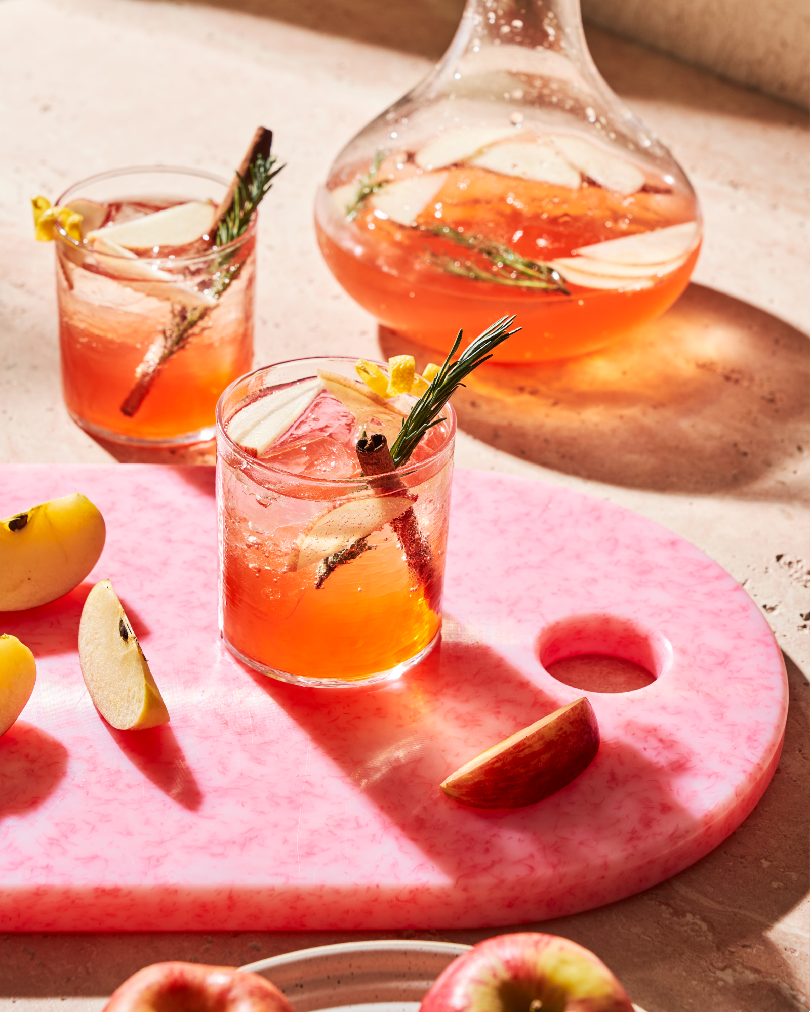 Two glasses of pink cocktails with garnishes on a Sasni Fairy Floss cutting board with apples.