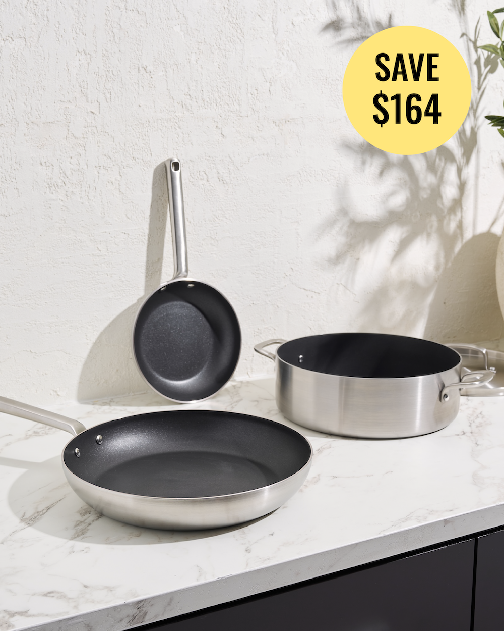 Three stainless steel cookware pieces on a white marble countertop against a textured white wall: a frying pan in front, a saucepan to the right, and a smaller pan leaning against the wall. A yellow circular badge in the top right reads 'SAVE $164'.