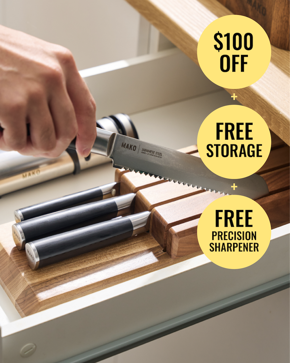 A hand inserts a Mako kitchen knife into a wooden drawer organizer. Three other Mako knives with black handles are already stored in the wooden slots.