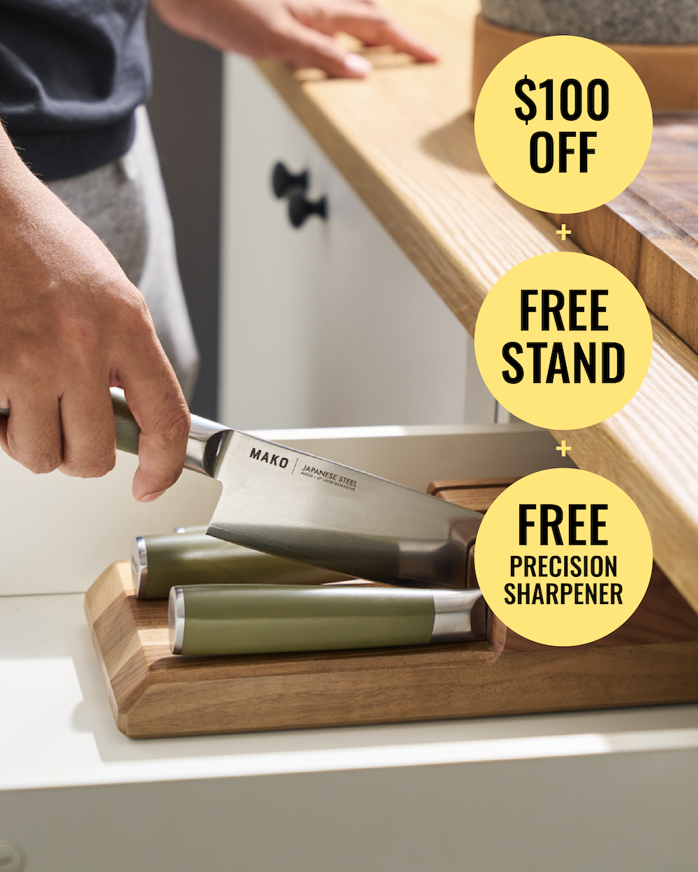 A person's hand holds a Mako professional chef knife over a wooden stand in a kitchen drawer. Two additional olive green-handled knives are visible in the stand.