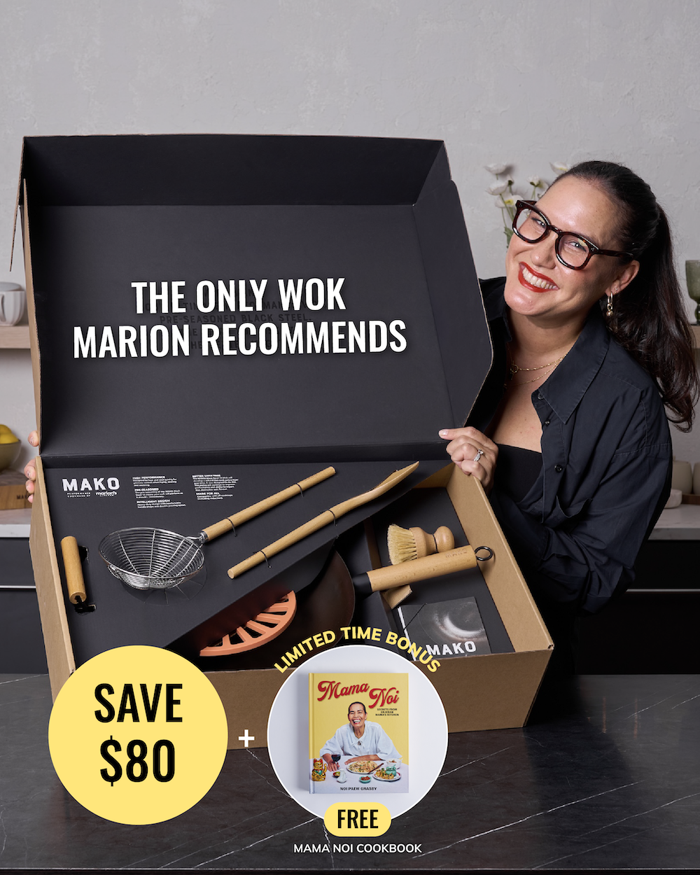 A woman in a black shirt and glasses smiles while opening a large Mako gift box set on a dark countertop. The open box contains cookware and accessories, including wooden utensils, a metal strainer, and a wok.