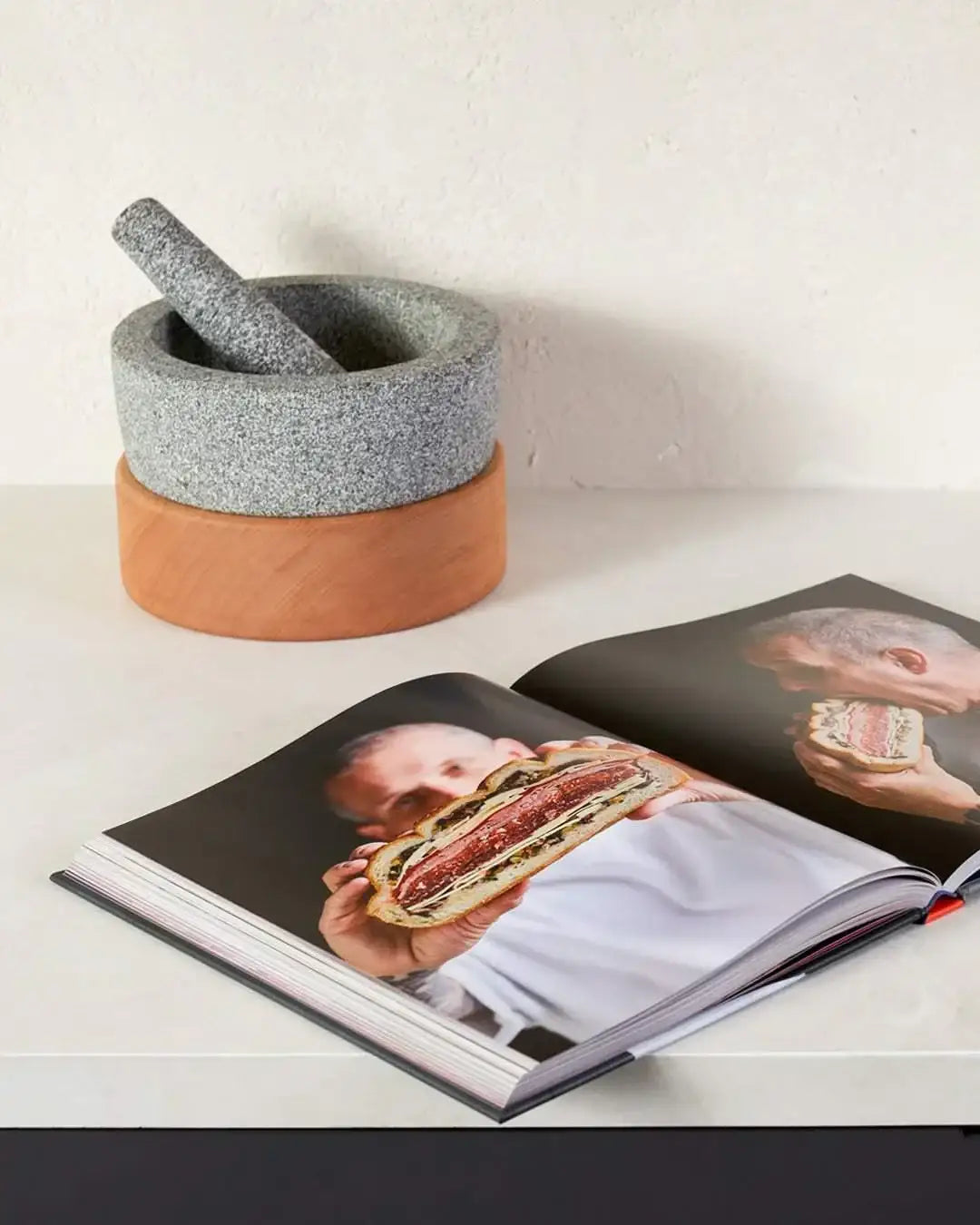 Granite mortar and pestle on wooden base next to open cookbook showing chef with sandwich