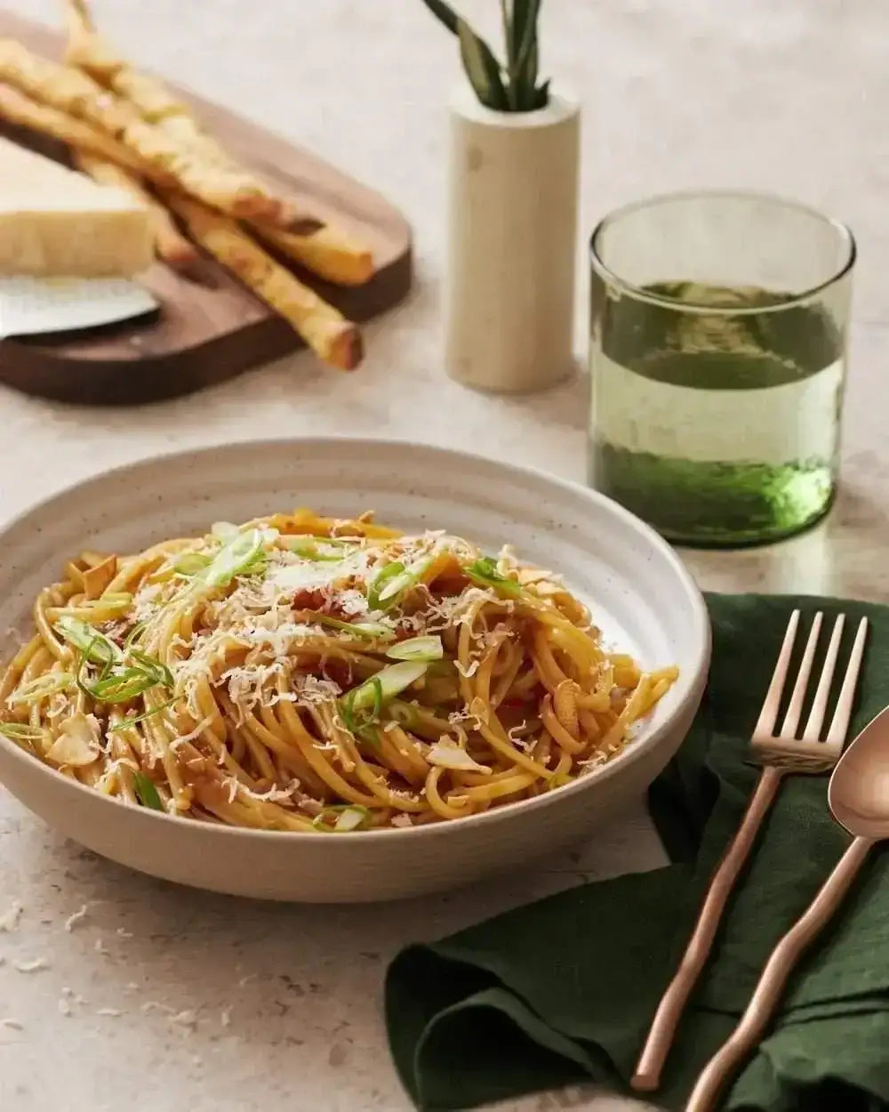 Asoke ceramic dinner plate with spaghetti topped with grated cheese and green onions, fork and spoon on green napkin, breadsticks and glass