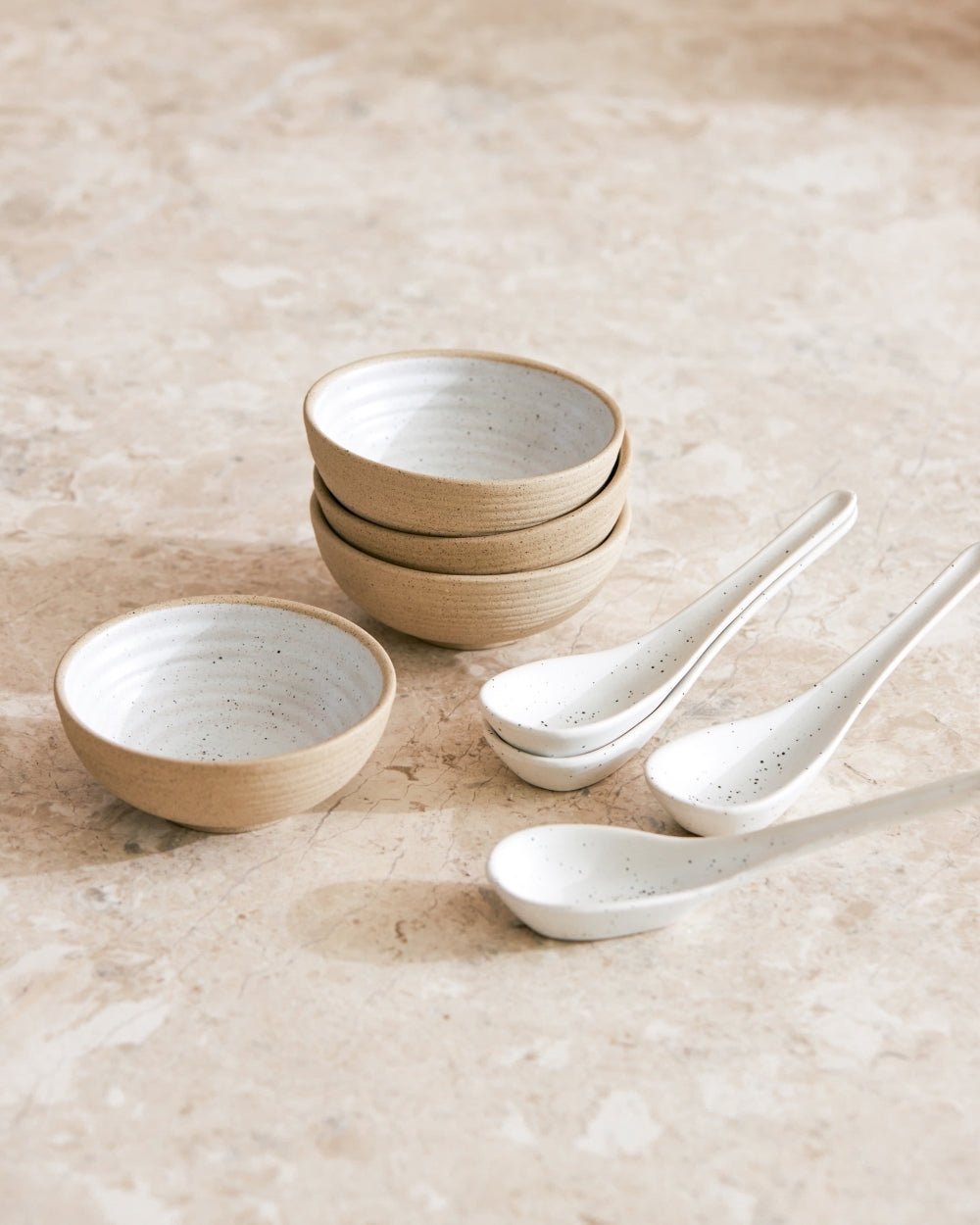 Set of four ceramic bowls and four ceramic spoons with natural, speckled finish on stone surface