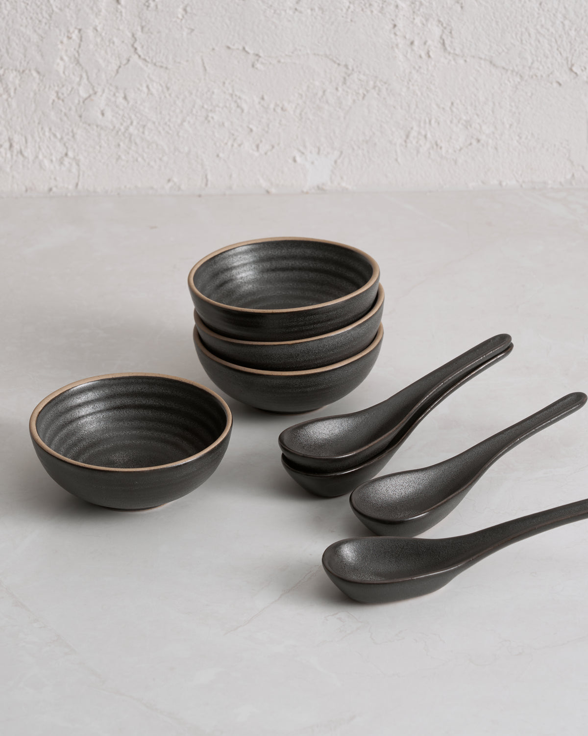 Charcoal stoneware bowls and ceramic soup spoons set on light textured surface