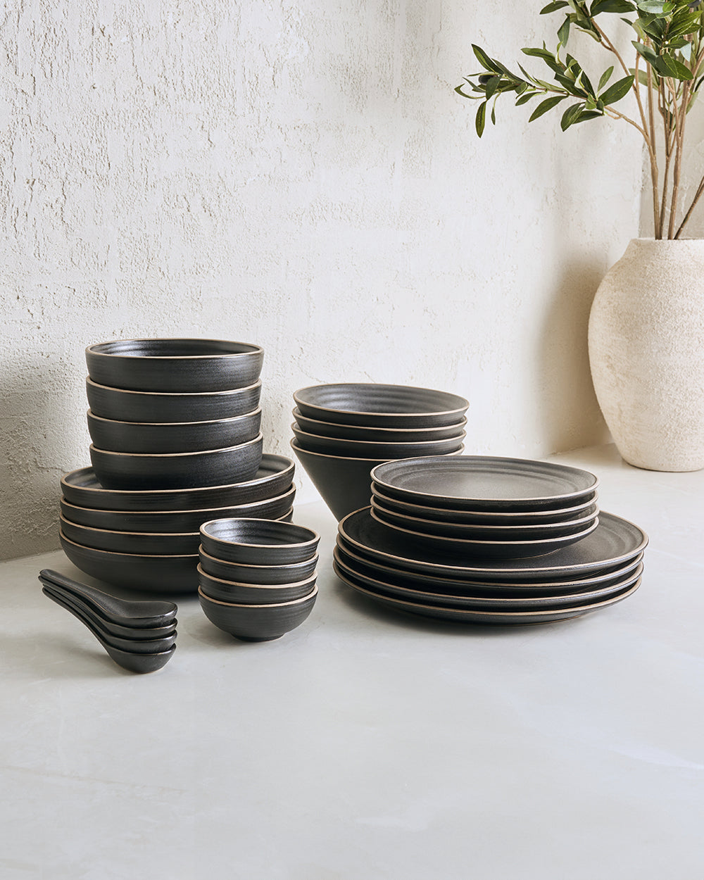 Anaya Charcoal Complete Dinnerware Set displayed elegantly against white textured wall, featuring stacked charcoal gray plates, bowls and spoons in various sizes, with a minimalist cream vase containing olive branches in background, showcasing modern ceramic dining collection