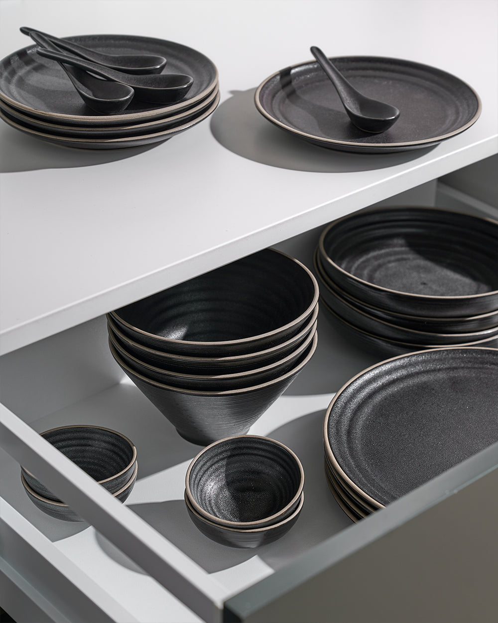 Anaya Charcoal Complete Dinnerware Set displayed on white shelving, featuring stacked charcoal gray plates, various sized bowls, and matching spoons arranged in drawer storage. Modern ceramic dinnerware with minimalist design and textured matte finish.