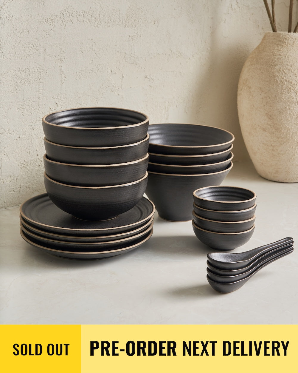 Anaya Charcoal Noodle Lovers Gift Set featuring stacked charcoal ceramic dinnerware including side plates, wide bowls, noodle bowls, mini bowls, and spoons, arranged on a white surface against a neutral textured wall with a beige ceramic vase in the background