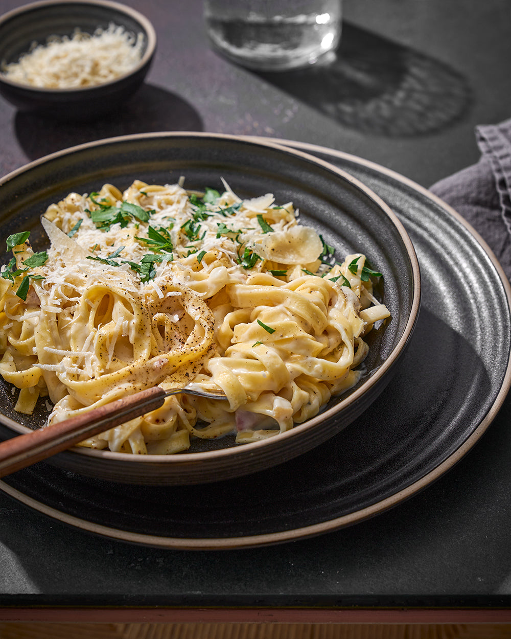 Anaya Charcoal Complete Dinnerware Set displayed with creamy fettuccine pasta garnished with parmesan cheese and fresh herbs, served in a matte black ceramic bowl on a matching charcoal dinner plate, with parmesan bowl and water glass in background