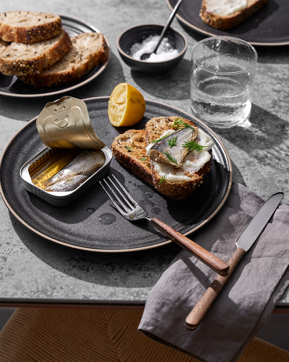 Anaya Charcoal Dinner Plate set of 4 showcasing a rustic table setting with seeded bread slices, canned sardines, and garnished open-faced sandwich served on a dark stoneware plate, accompanied by cutlery with wooden handles and a glass of water on a grey surface.