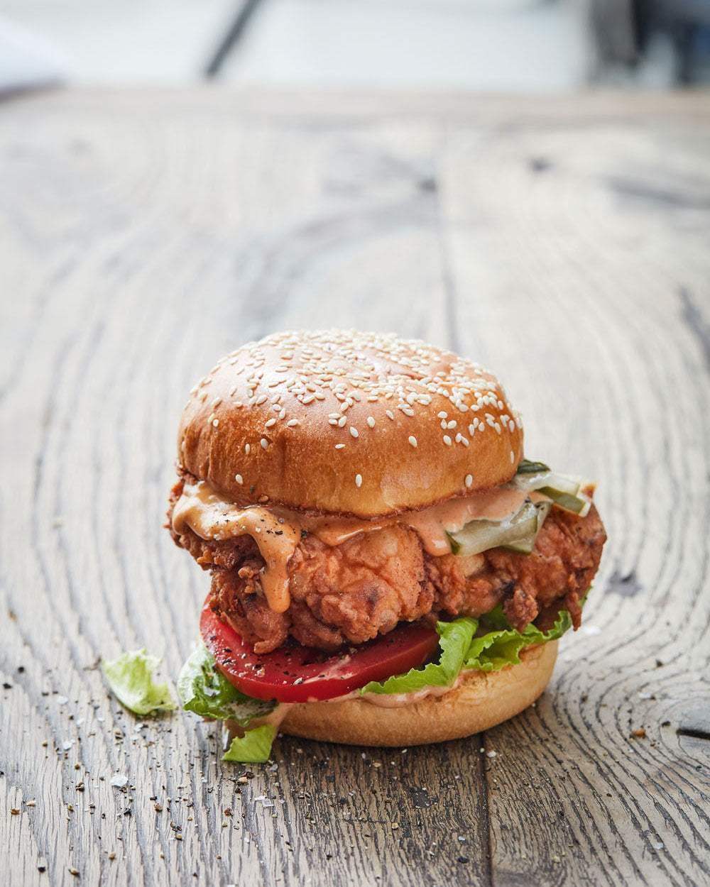 Crispy fried chicken sandwich with lettuce, tomato, pickles, and spicy sauce on sesame bun on rustic wood table