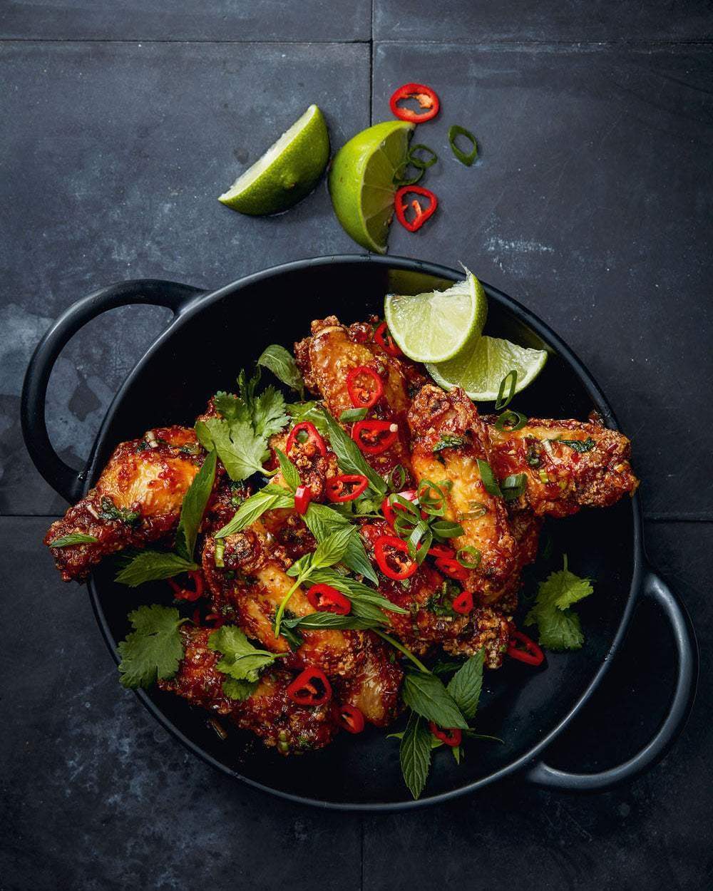 Fried chicken wings garnished with red chili slices, fresh herbs, and lime wedges in a black pan