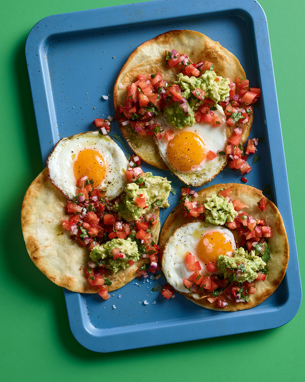 Three tostadas topped with sunny side eggs, guacamole, and tomato salsa on a blue tray