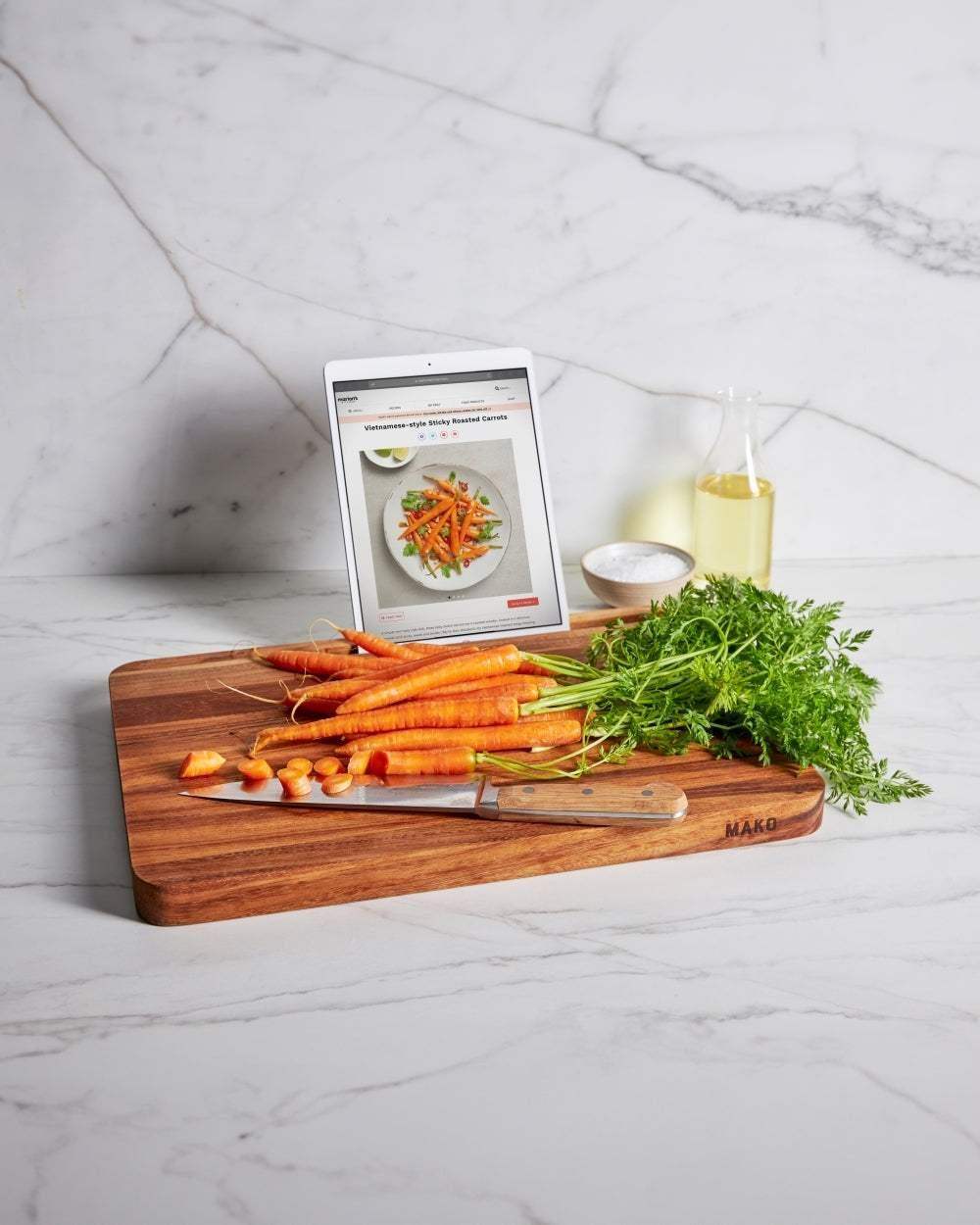 Mako Prestige wooden chopping board with fresh carrots, knife, salt bowl, oil bottle, and tablet showing a carrot recipe