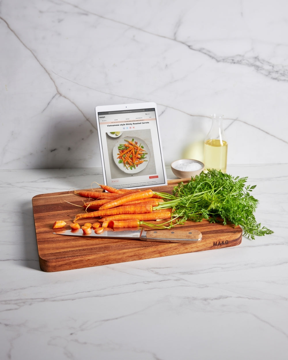 Wooden chopping board with fresh carrots, knife, and recipe on a tablet in kitchen setting