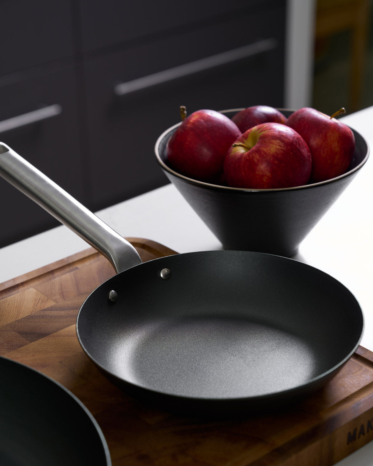 Black steel frying pan with stainless handle on cutting board, bowl of red apples in background