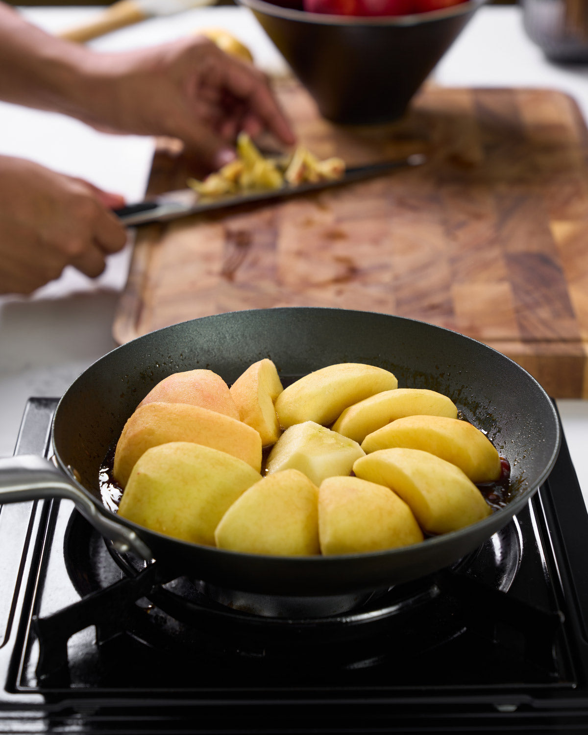 Black steel frying pan on stove with sliced apples cooking, person chopping fruit in background