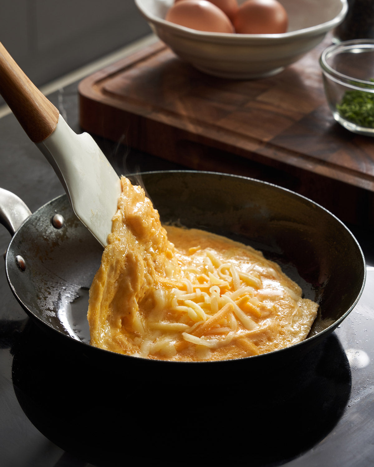 Cheese omelette cooking in black steel frying pan on stovetop, eggs and herbs in background