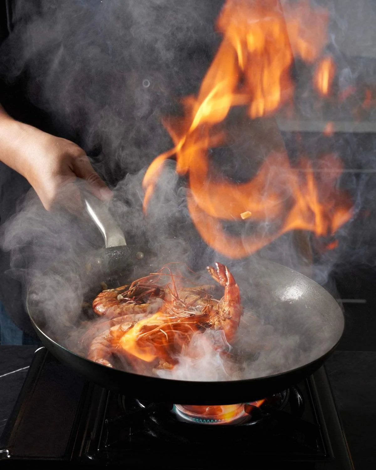 Black steel frying pan on stovetop with prawns, visible flames and steam rising during cooking