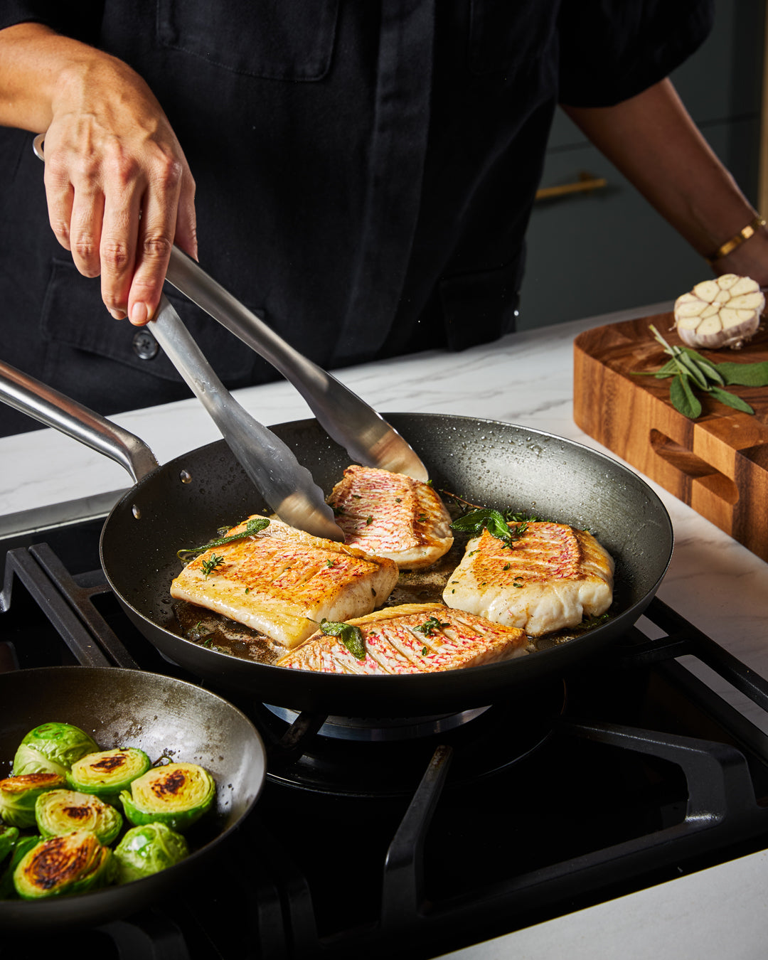 Person cooking fish fillets in a black steel frying pan on stovetop with seared Brussels sprouts nearby.