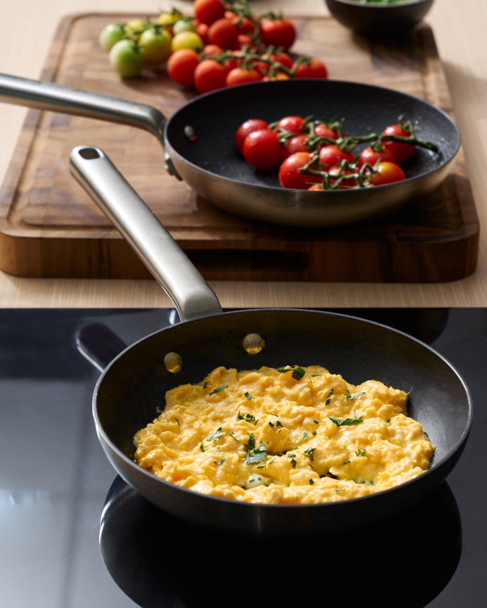 Two non-stick frying pans, one with scrambled eggs and one with cherry tomatoes, on a kitchen counter.