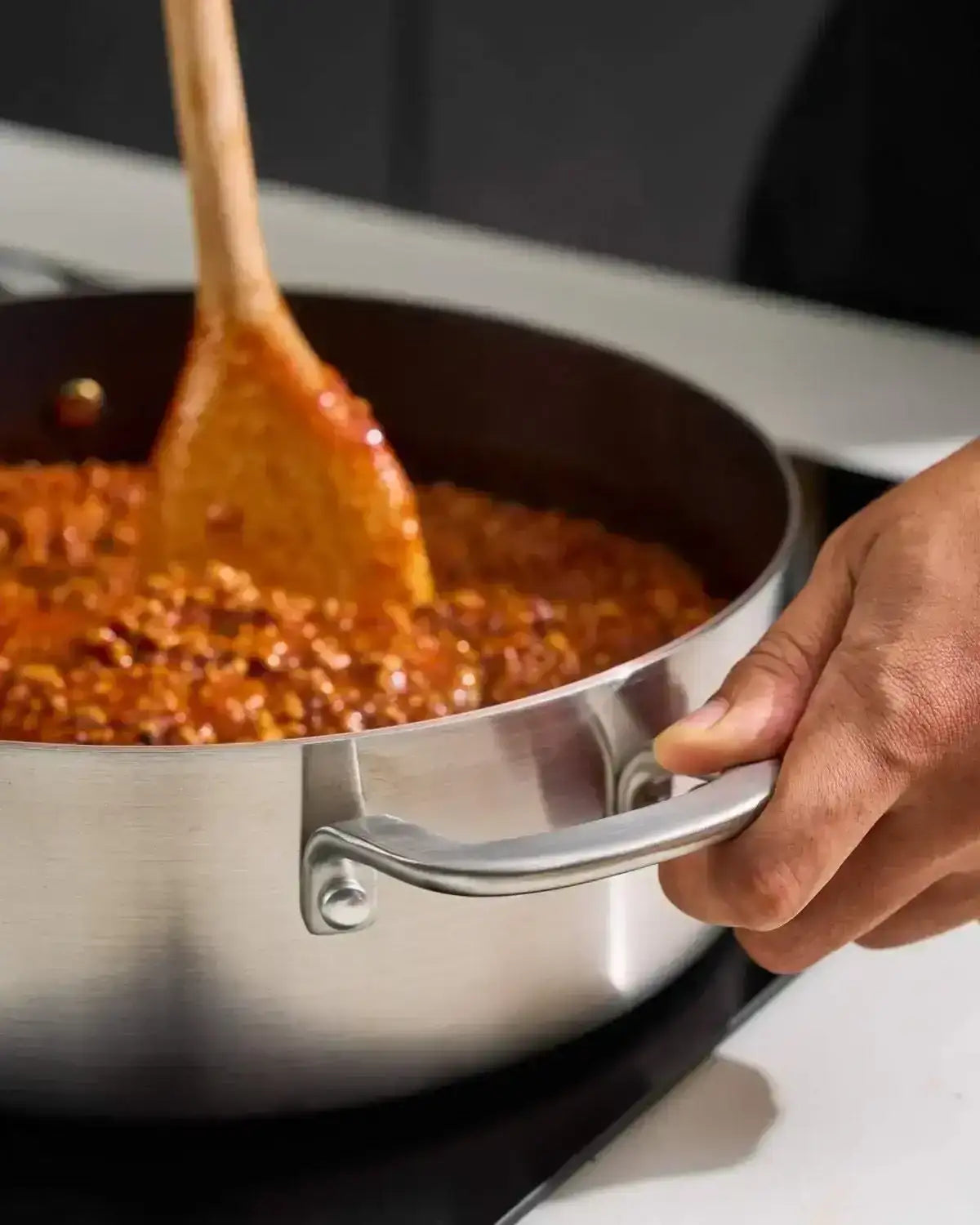 Close-up of hand holding steel handle of Mako stainless steel pan cooking tomato sauce with wooden spatula