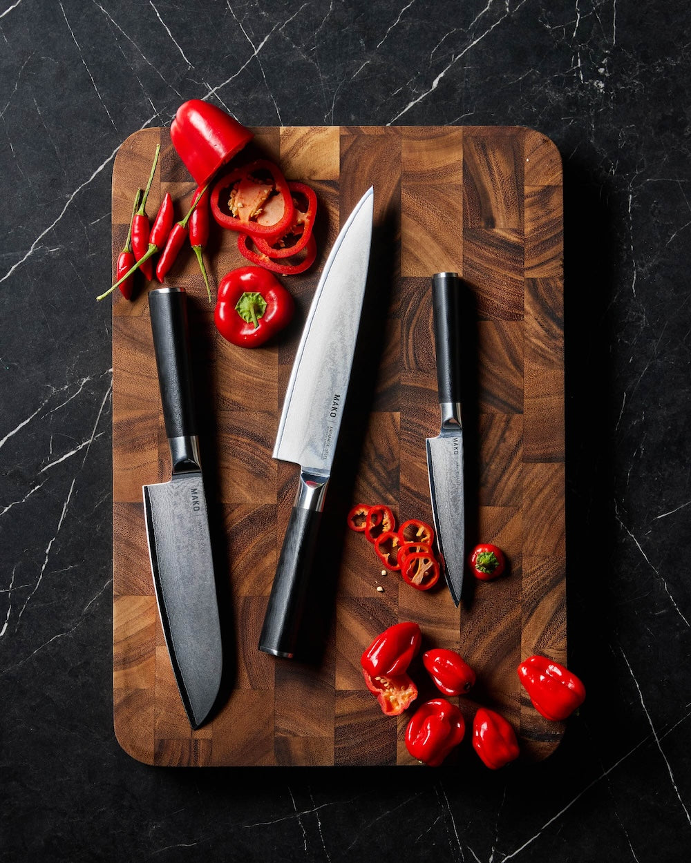 Three kitchen knives on wooden cutting board with sliced red peppers on black marble countertop