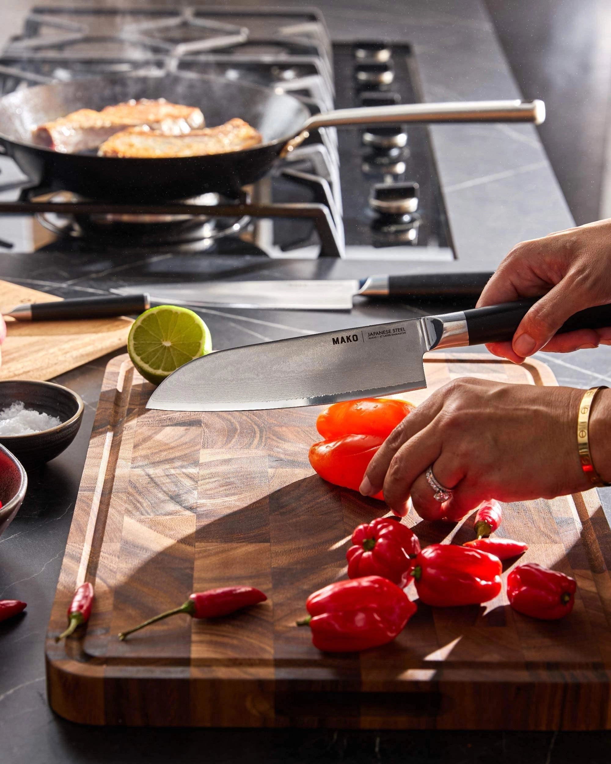 Slicing tomato with Japanese steel chef knife on wooden cutting board, peppers and lime nearby, kitchen in background.