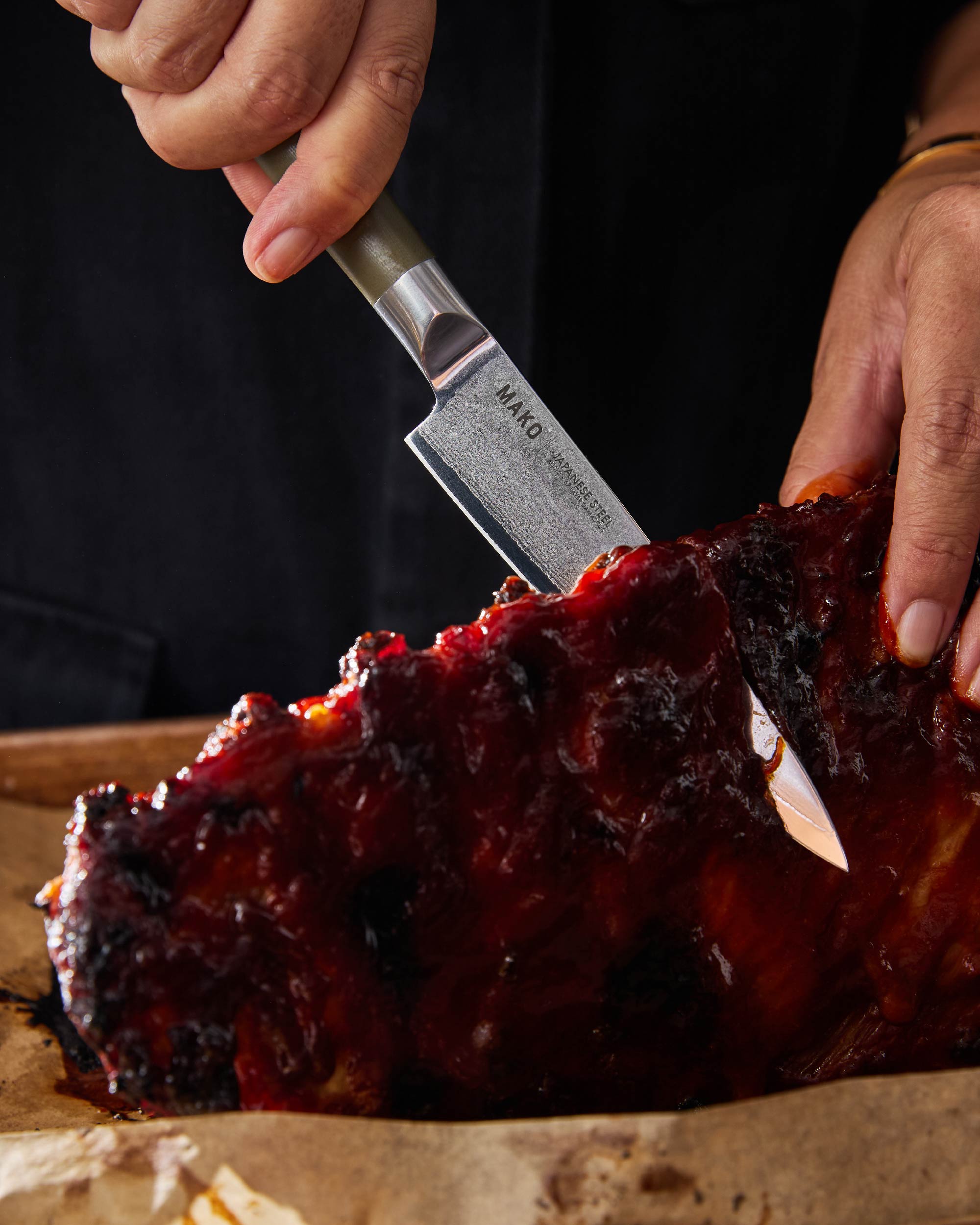 Person slicing barbecued ribs with Japanese steel kitchen knife, olive green handle, close-up