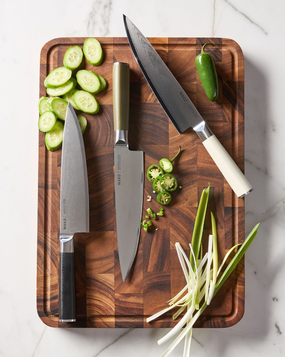 Three Japanese steel chef knives with assorted handles on a wooden cutting board with sliced vegetables.