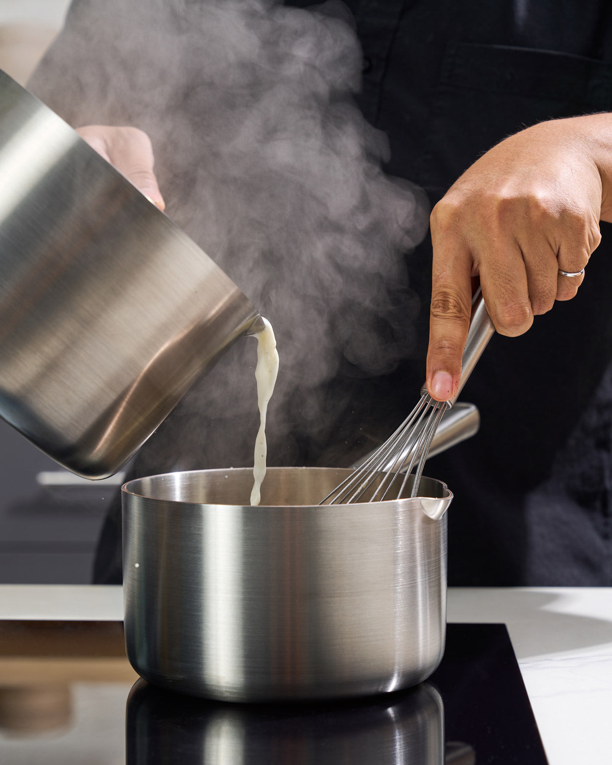 Stainless steel saucepan on cooktop with whisk and liquid being poured, kitchen scene