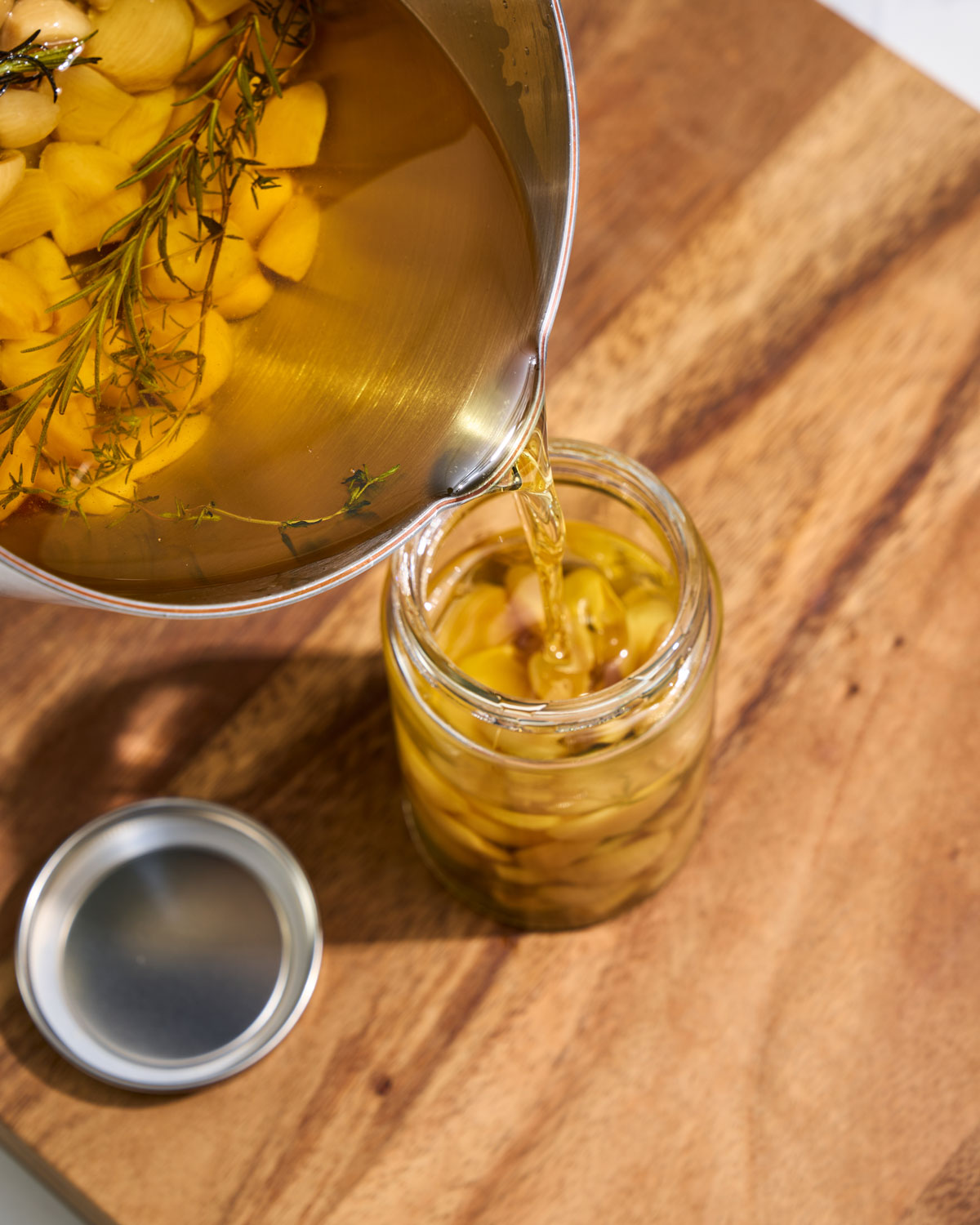 Stainless steel saucepan pouring herb and garlic oil into glass jar on wood board
