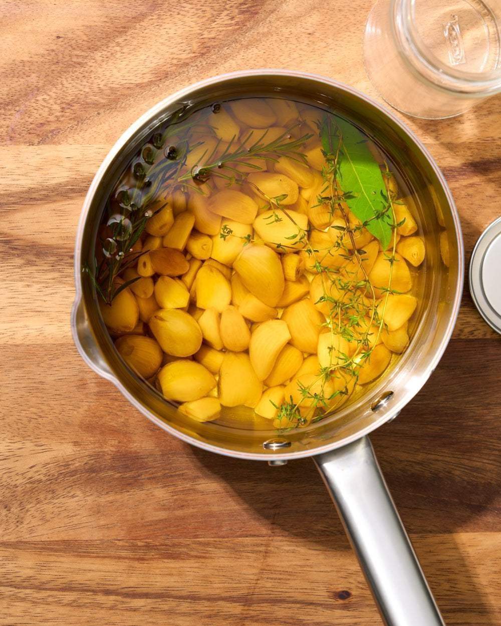 Stainless steel saucepan with peeled garlic cloves and fresh herbs soaking in liquid on wooden surface