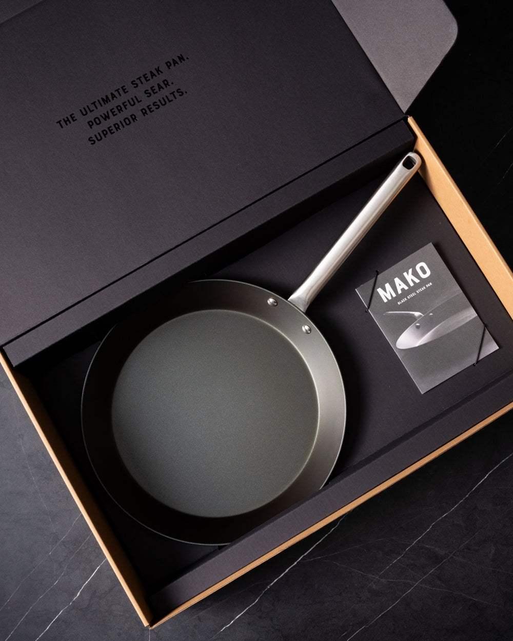 Black steel steak pan with long handle in gift box on dark surface, product booklet included.