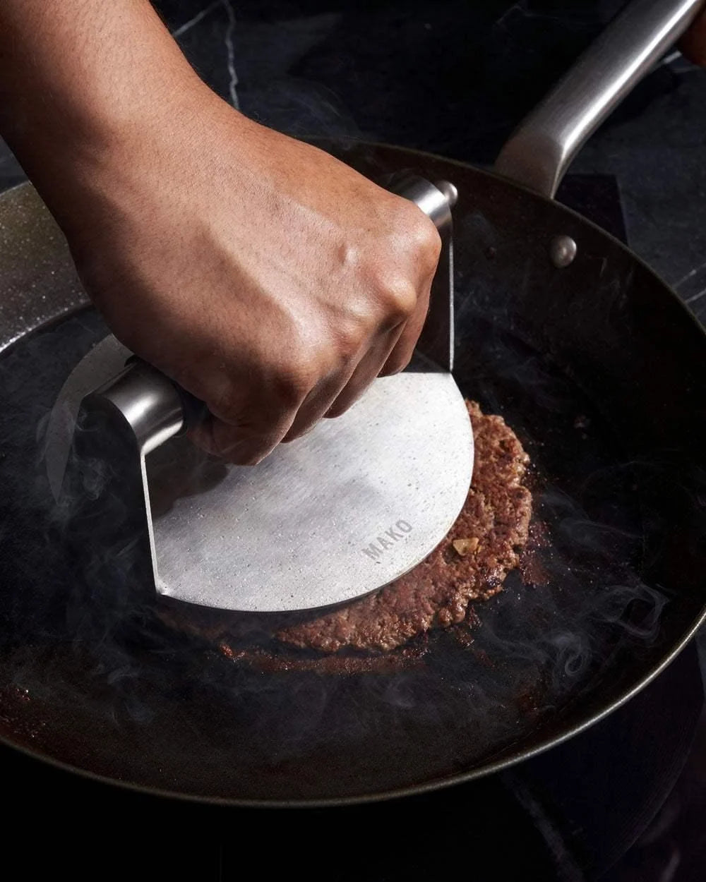 Hand pressing burger patty with metal press in black steel pan on stovetop