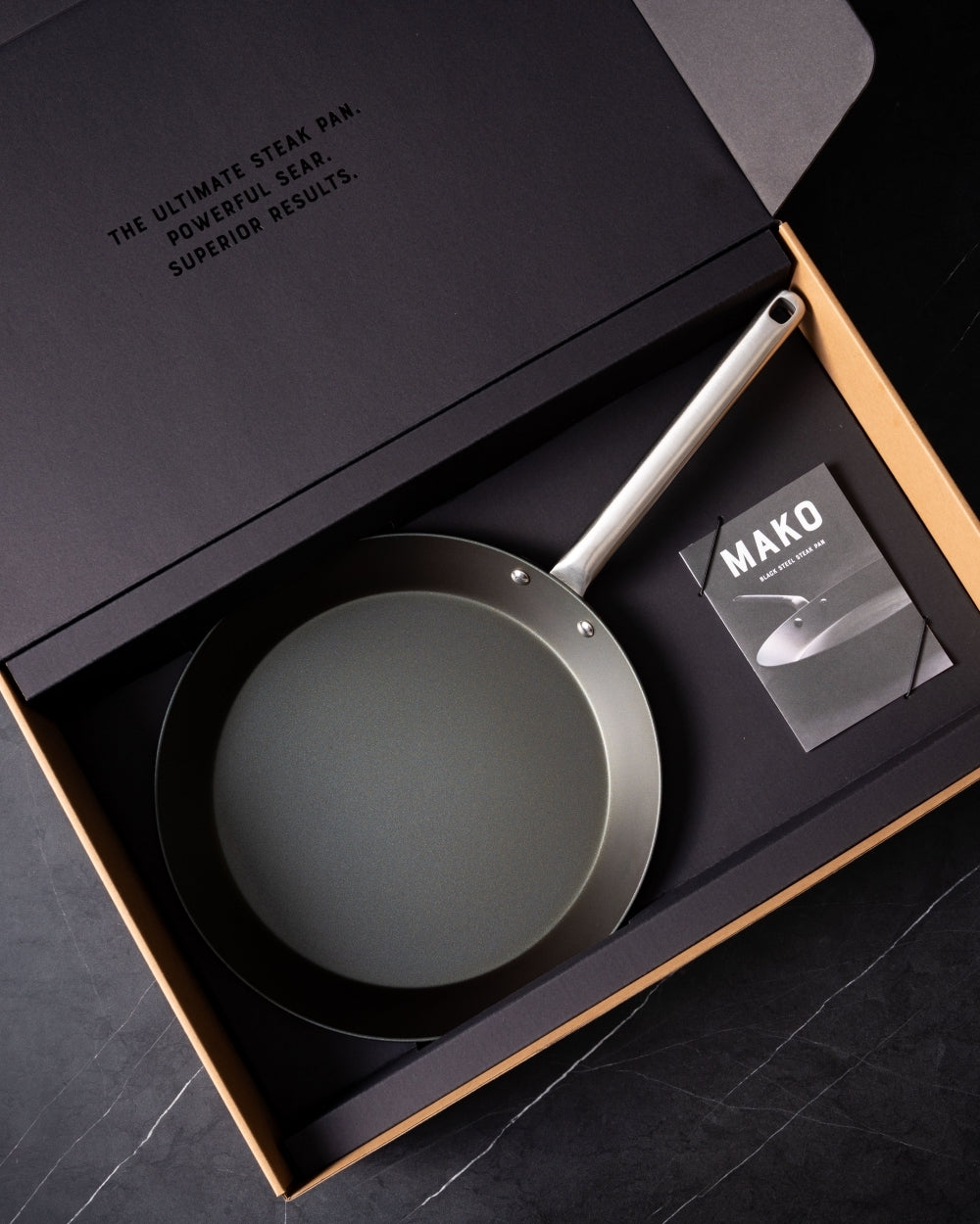 Black steel frying pan with metal handle in gift box, MAKO booklet included, on dark surface.
