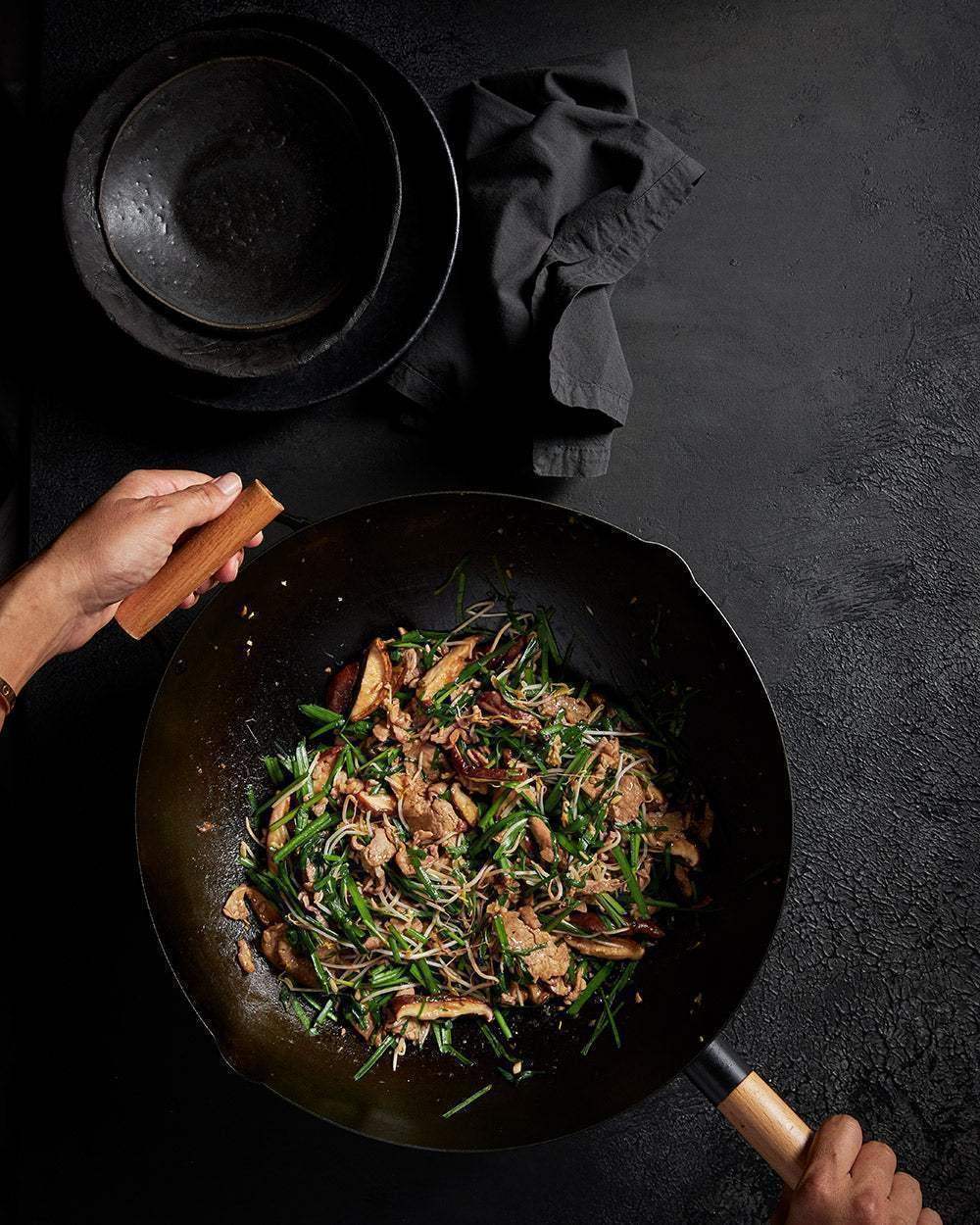 Marion Grasby’s Mako wok with wooden handle cooking stir-fry with mushrooms and greens on charcoal background