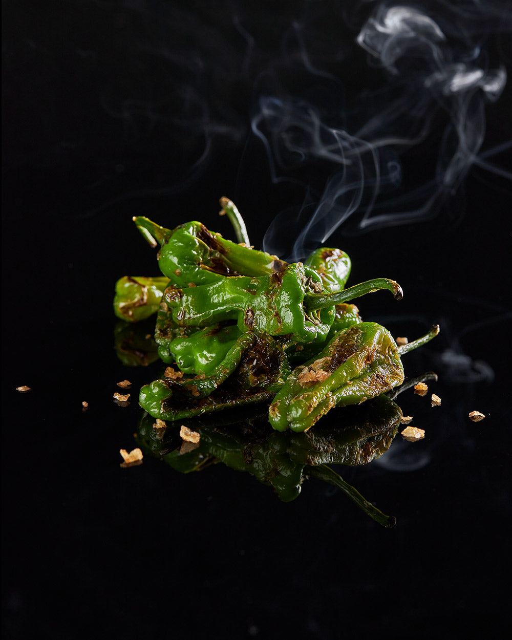 Charred green shishito peppers with flakes and rising steam on a glossy black background.